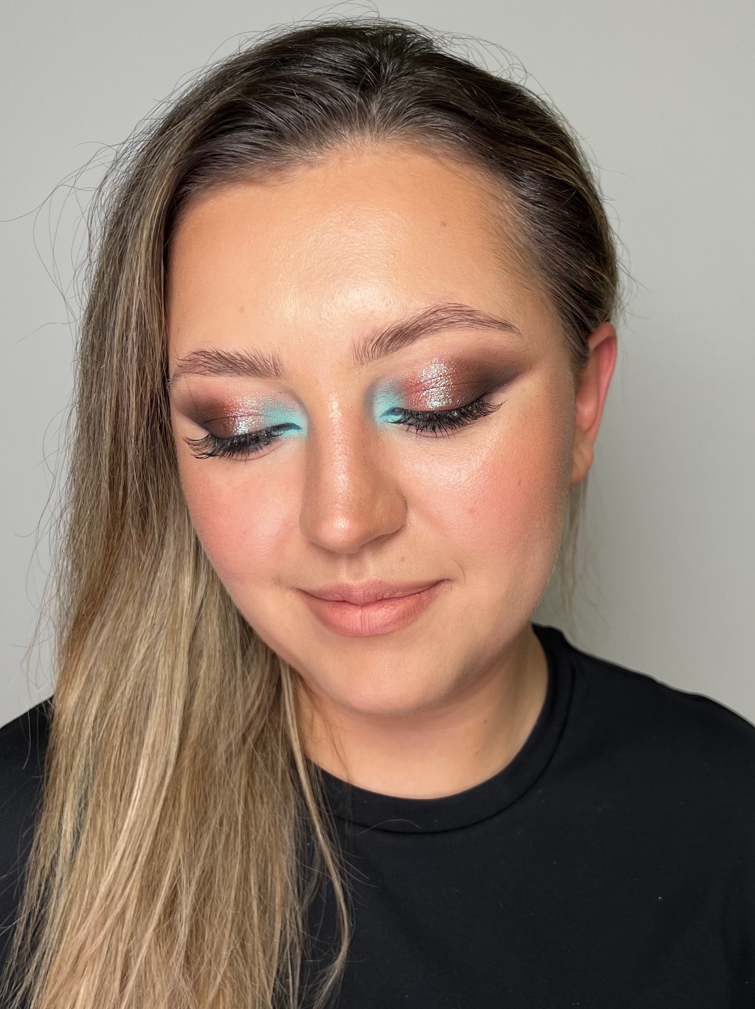 Portret kobiety z zamkniętymi oczami, prezentujący intensywny, błyszczący makijaż oczu w odcieniach brązu i turkusu, z wyraźnie podkreślonymi rzęsami.