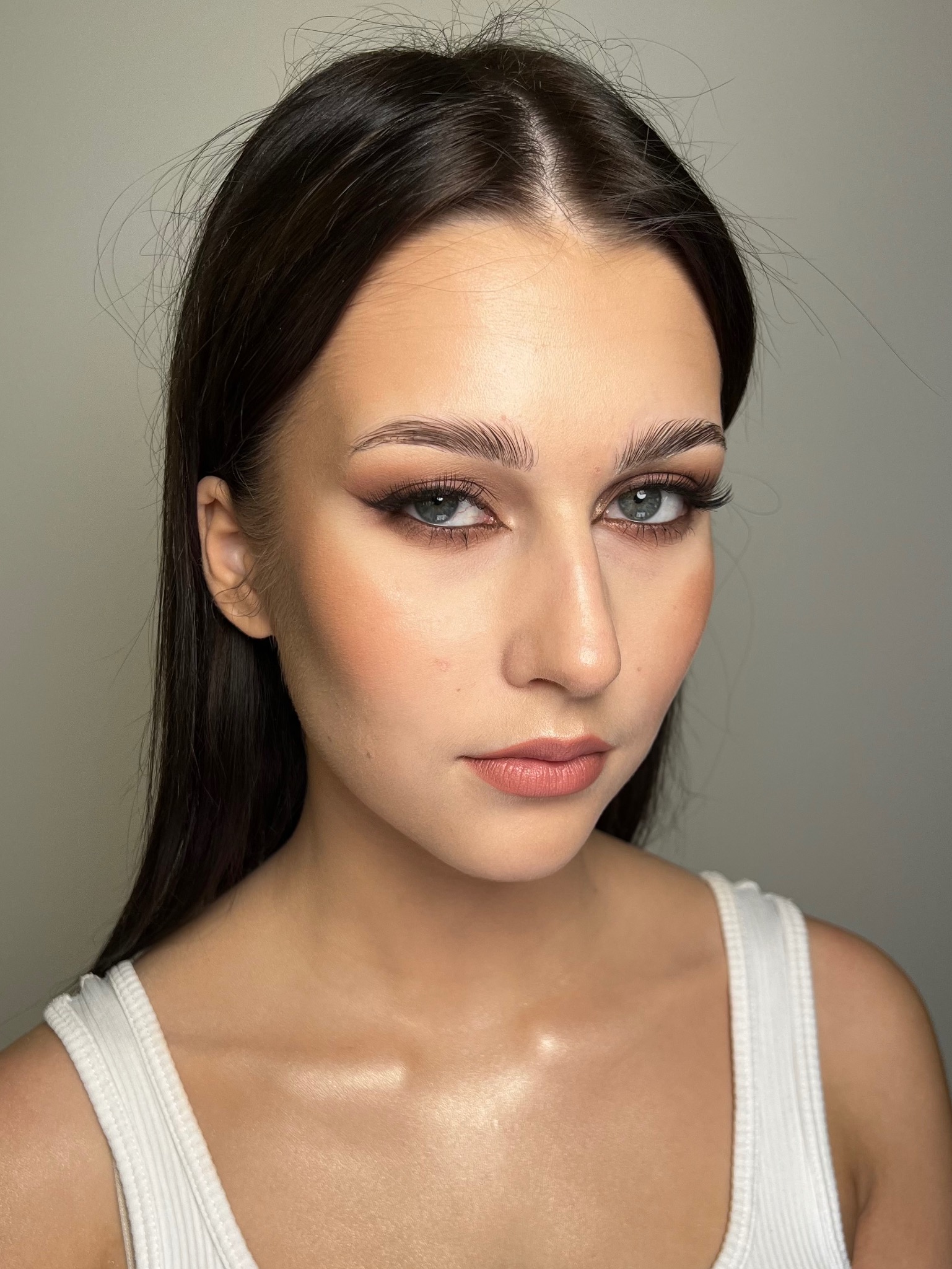 Portret kobiety z precyzyjnym makijażem oczu w stylu 'kocie oko', podkreślonymi brwiami i delikatnym makijażem ust, na tle jasnej ściany.