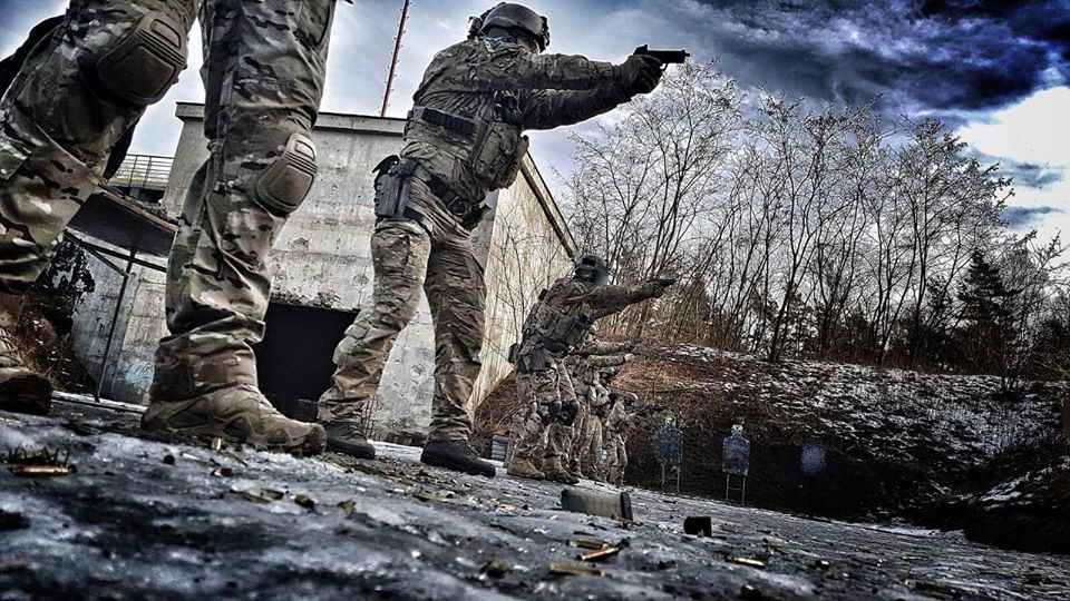 Szkolenie taktyczne w Warszawie: grupa umundurowanych mężczyzn celuje z broni krótkiej do tarcz na poligonie, widoczne łuski naboi i betonowy budynek w tle.