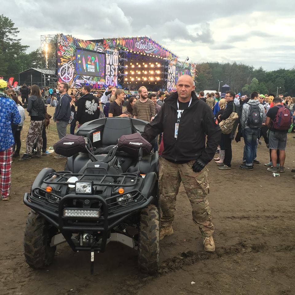 Mężczyzna w odzieży taktycznej stoi obok quada na tle sceny festiwalowej z tłumem ludzi.