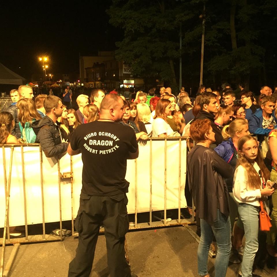 Ochroniarz w czarnym uniformie z napisem 'Biuro Ochrony Dragon Robert Pietrzyk' stoi tyłem do kamery, pilnując porządku za metalowymi barierkami podczas nocnego wydarzenia plenerowego z dużym...