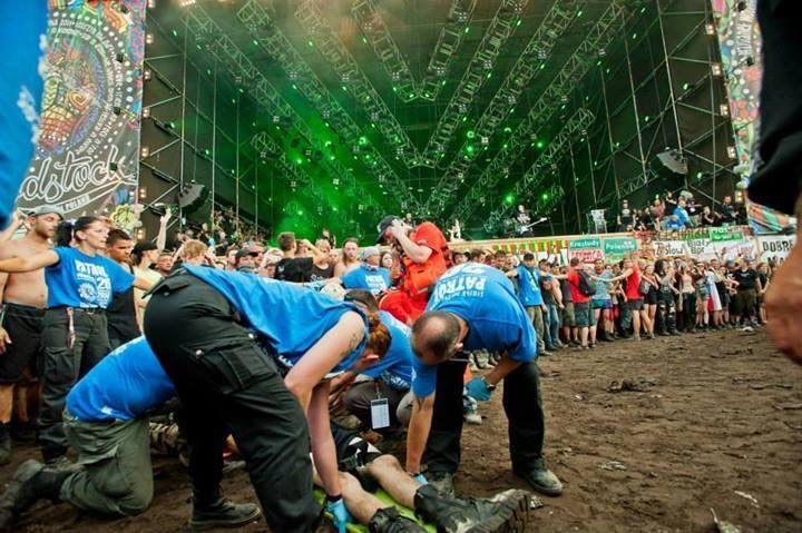Ratownicy medyczni udzielają pomocy poszkodowanemu na festiwalu muzycznym, na tle sceny z oświetleniem i tłumu ludzi.