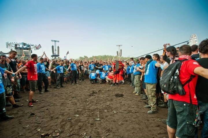 Grupa ratowników medycznych w czerwonych strojach udziela pomocy osobie na ziemi podczas masowej imprezy plenerowej. Wokół nich tłum ludzi w niebieskich koszulkach tworzy barierę, trzymając się za...