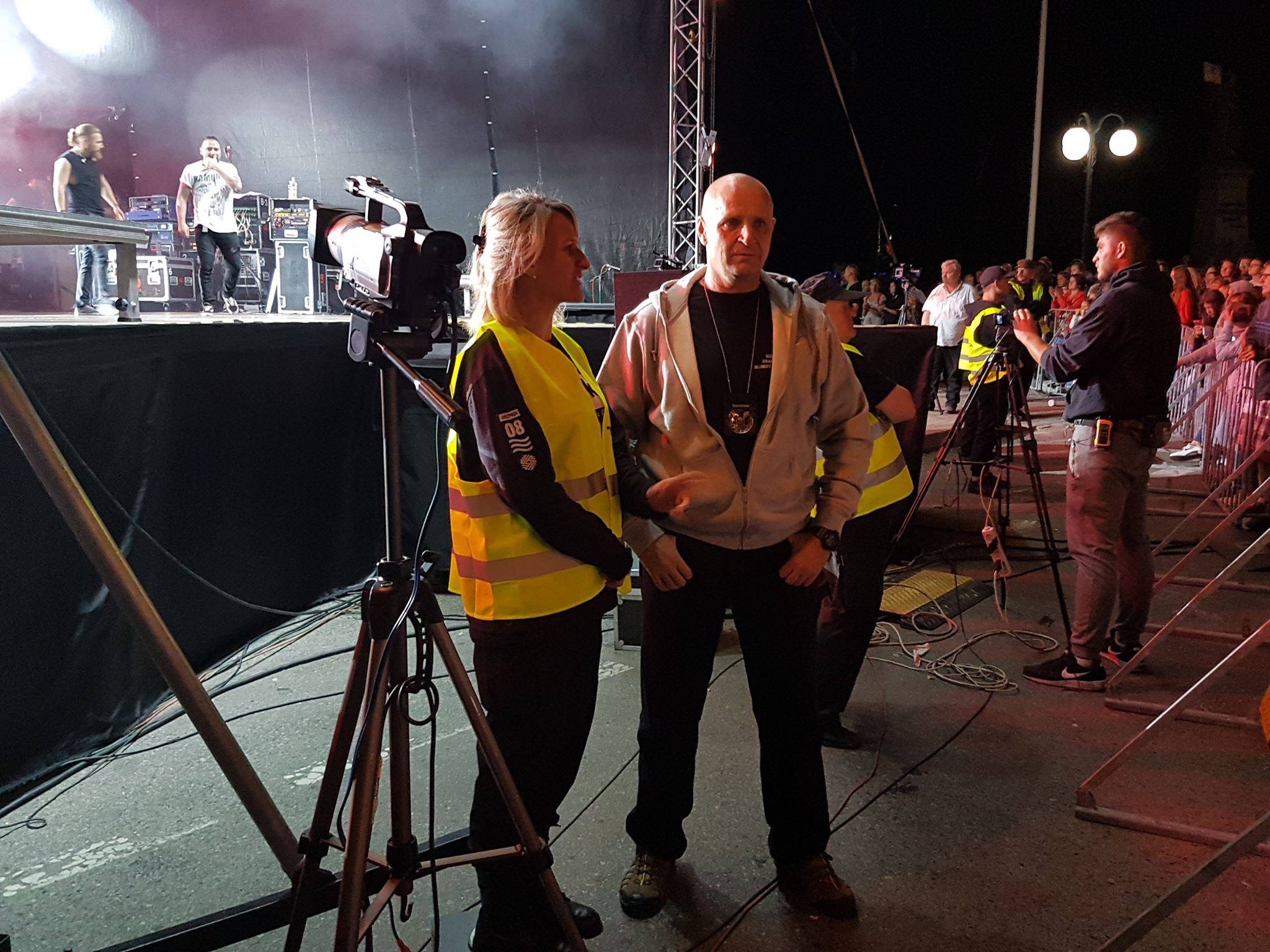 Ochrona imprezy masowej: mężczyzna i kobieta w kamizelkach odblaskowych stoją przed kamerą, w tle scena z występem i publiczność.