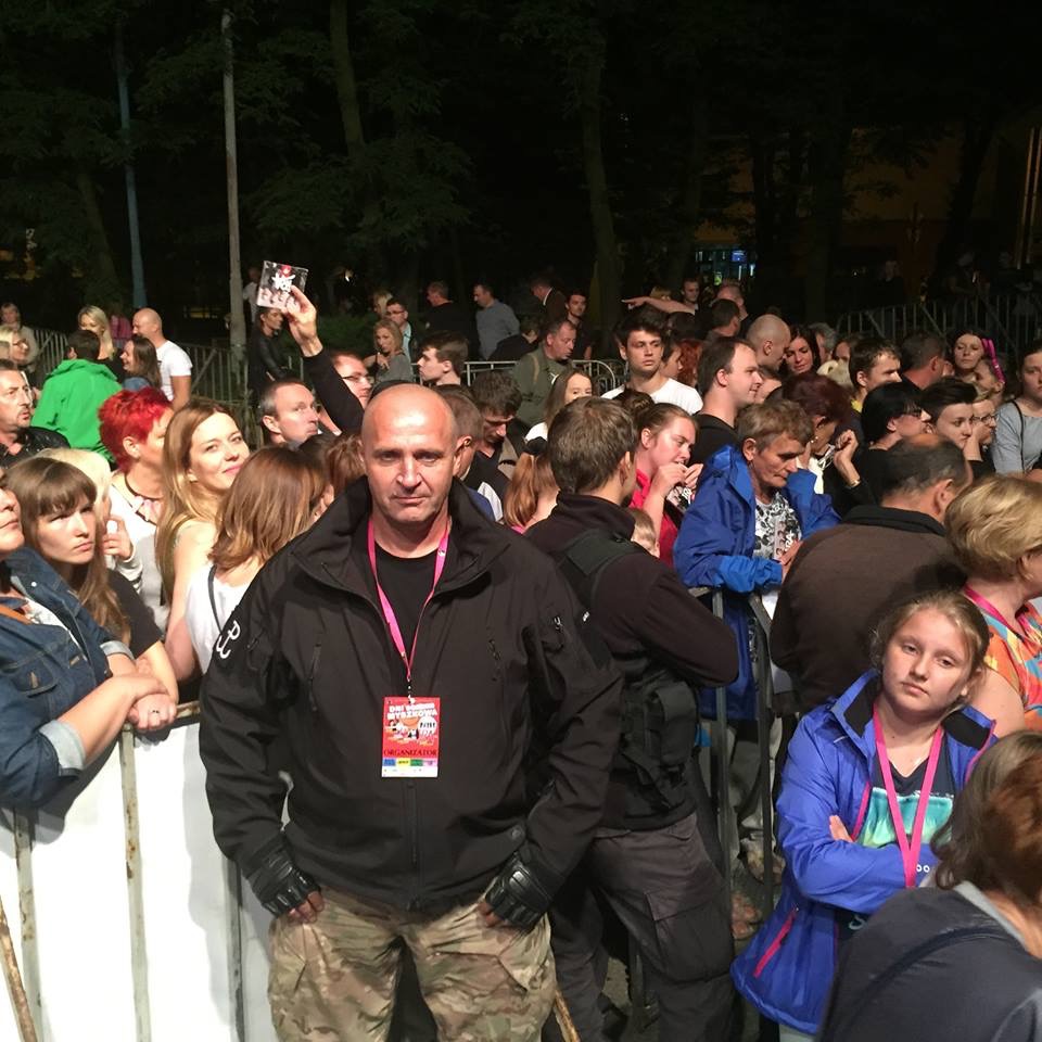 Mężczyzna w czarnej kurtce i spodniach moro stoi przed barierkami na tle tłumu ludzi podczas wydarzenia plenerowego, na szyi ma identyfikator.