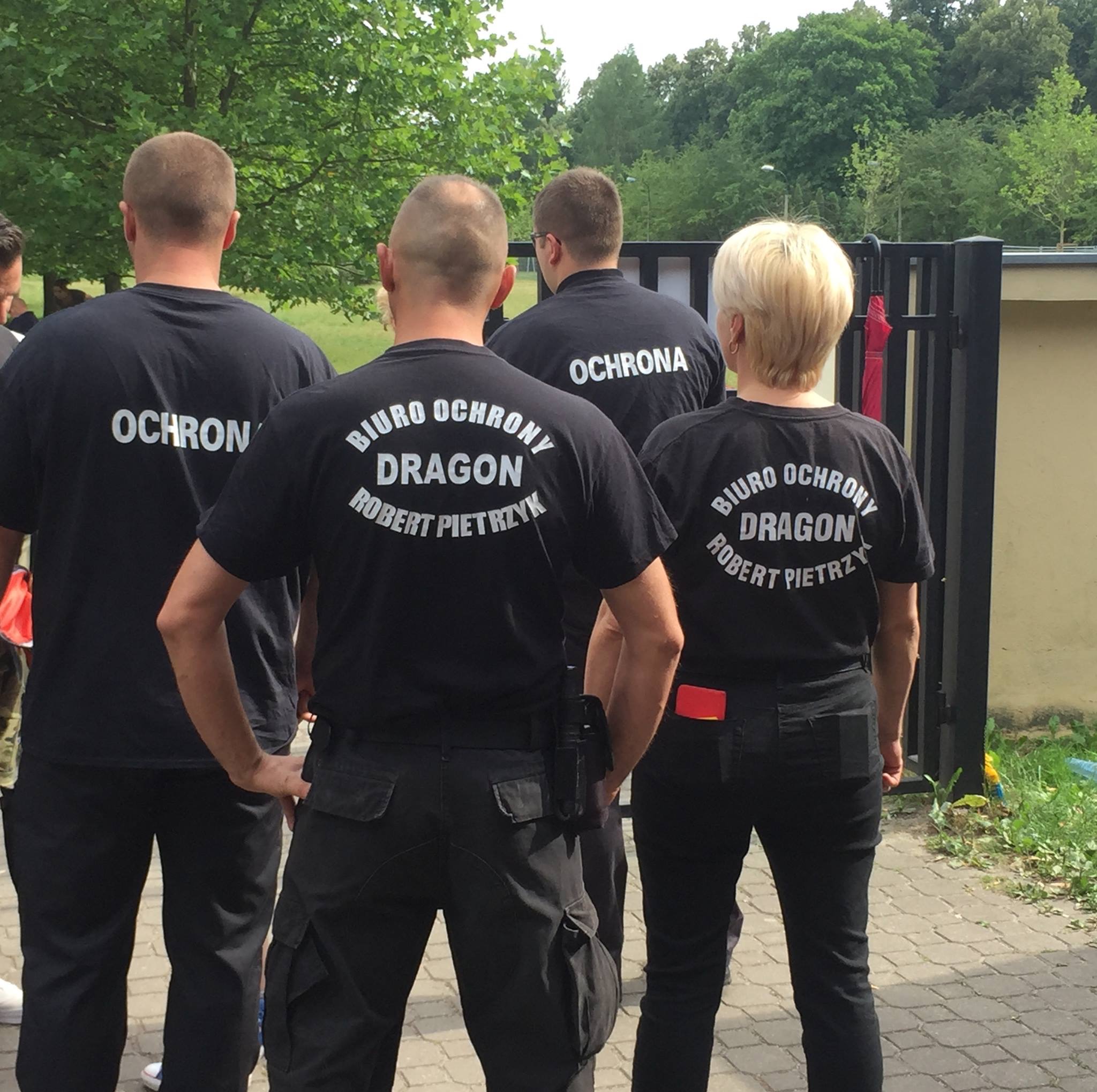 Grupa osób z tyłu, ubrana w czarne koszulki z napisem 'Biuro Ochrony Dragon Robert Pietrzyk' oraz 'Ochrona', stojąca na zewnątrz, prawdopodobnie podczas wykonywania obowiązków.
