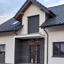 Nowoczesny dom jednorodzinny z białą elewacją, grafitową dachówką i stolarką okienną, z częścią frontową wykończoną cegłą klinkierową w odcieniach szarości oraz młodym drzewkiem o purpurowych...