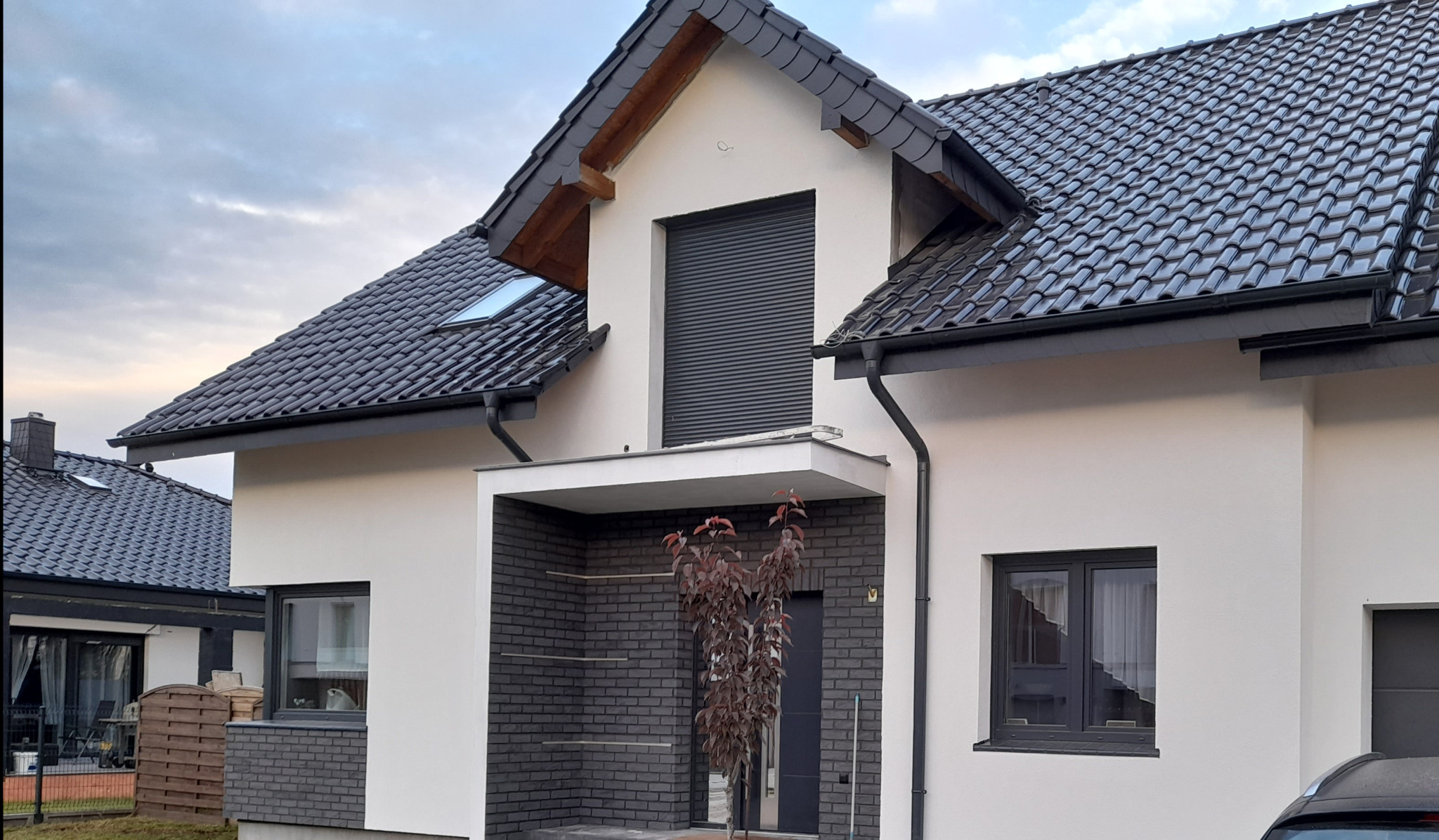 Nowoczesny dom jednorodzinny z białą elewacją, grafitową dachówką i stolarką okienną, z częścią frontową wykończoną cegłą klinkierową w odcieniach szarości oraz młodym drzewkiem o purpurowych...