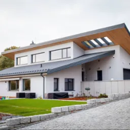 Nowoczesny dom jednorodzinny z płaskim dachem i drewnianym wykończeniem okapu oraz oknami dachowymi, widok z zewnątrz na elewację i zagospodarowany teren z kostką brukową i trawnikiem