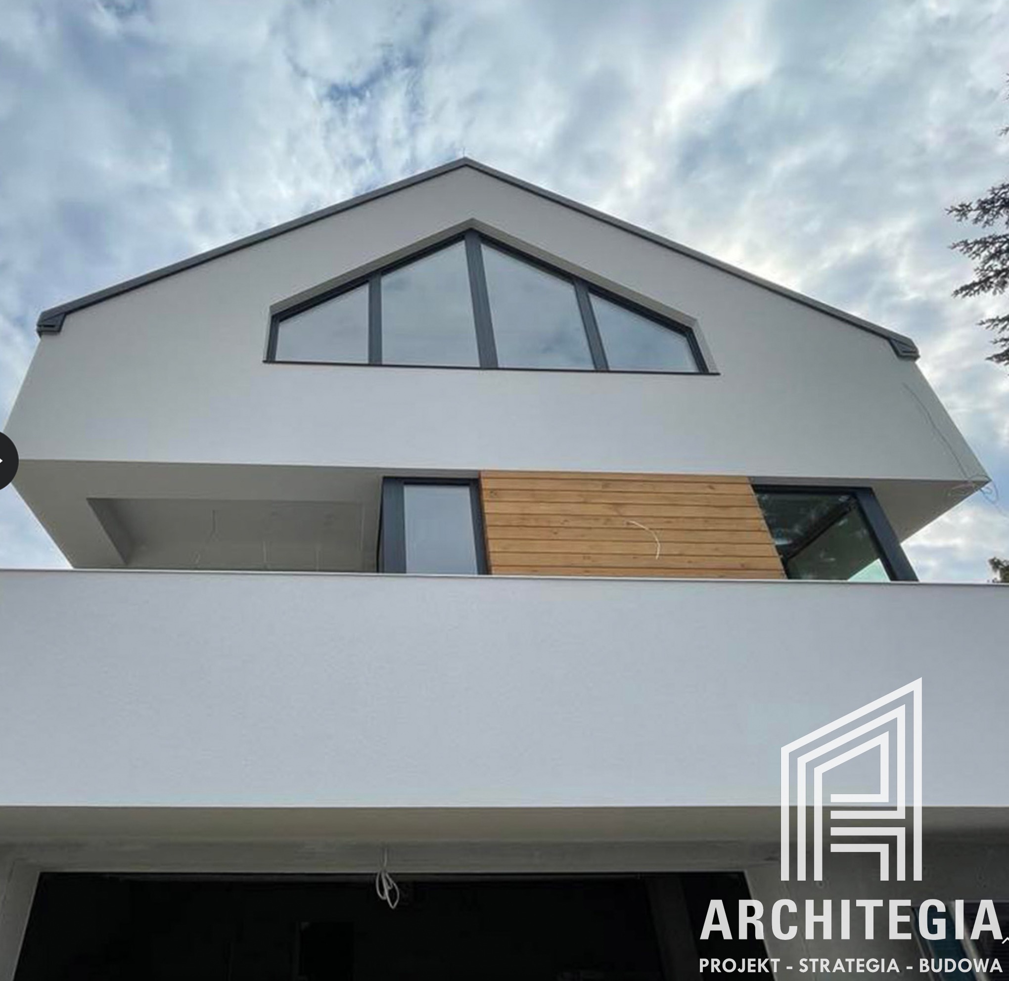 Nowoczesny dwupiętrowy dom z płaskim dachem i dużymi oknami, wykończony jasnym tynkiem i elementami z drewnianych paneli, widoczny z dołu w pochmurny dzień. Na dole widoczne logo firmy...