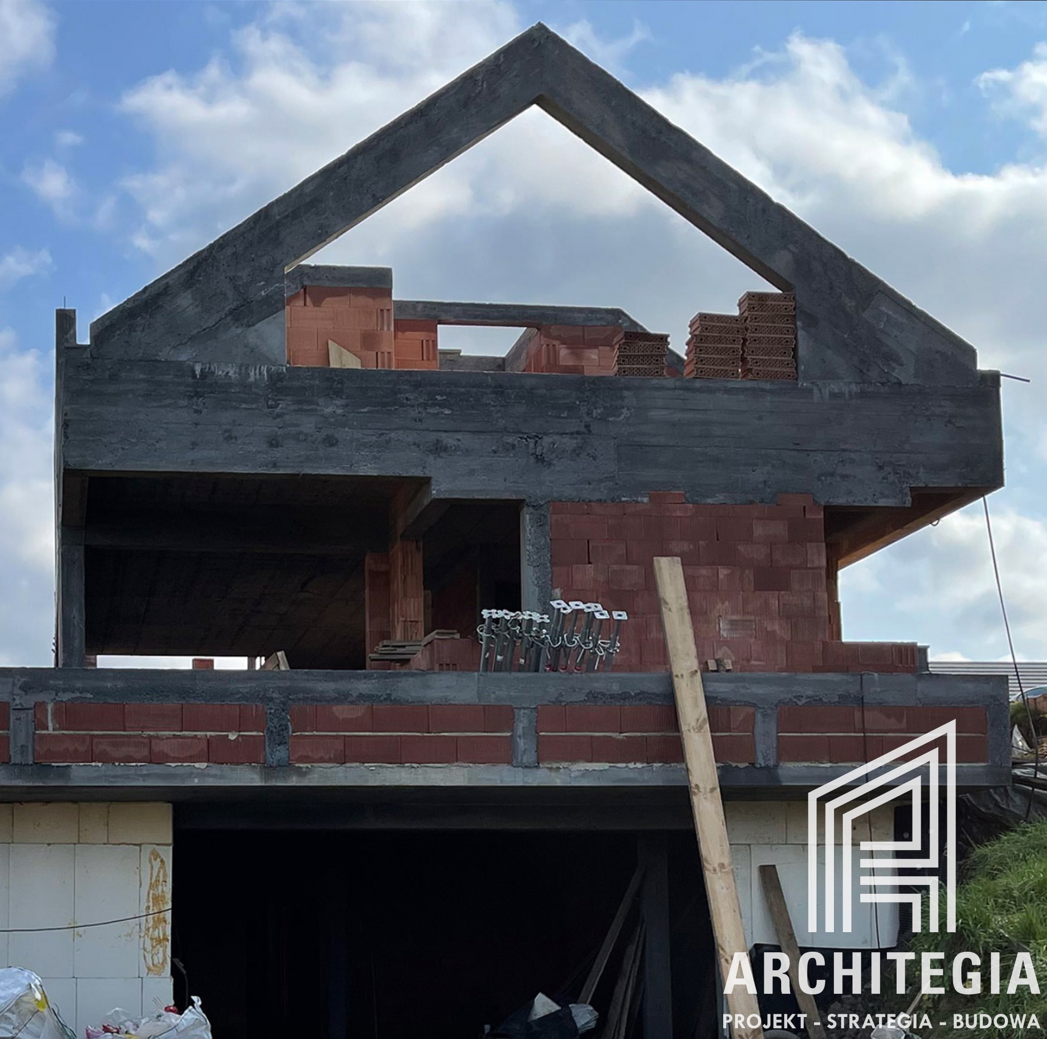 Surowy stan domu z czerwonej cegły z widocznym szalunkiem betonowym oraz logo firmy Architegia w prawym dolnym rogu.