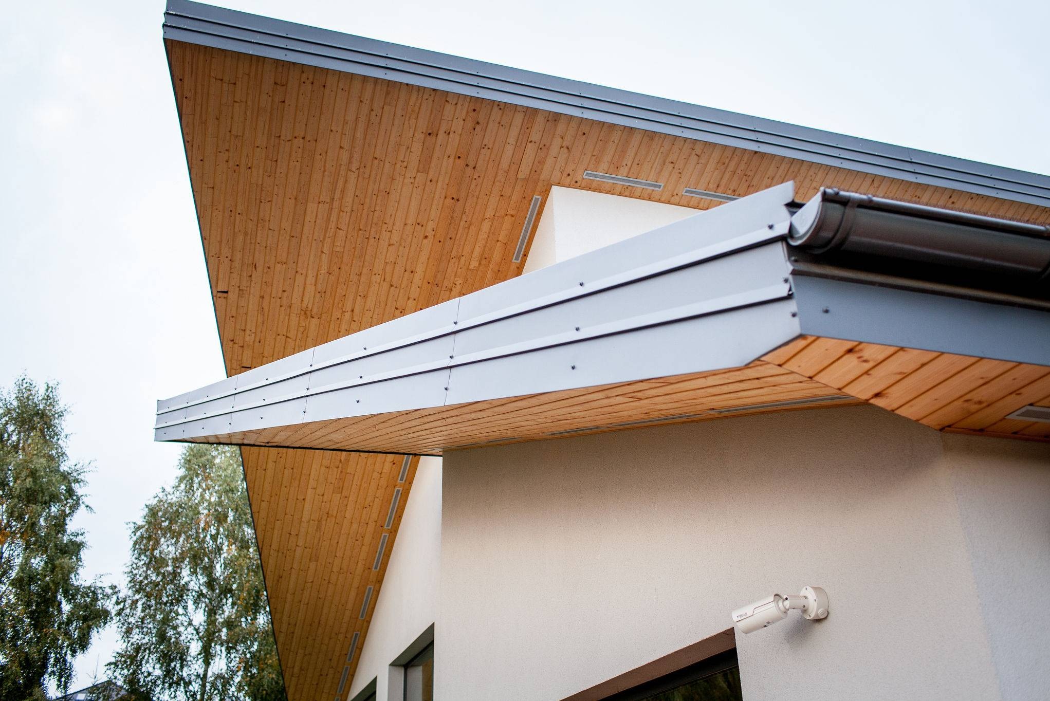 Nowoczesny dom z charakterystycznym drewnianym podbitkiem dachowym i metalową obróbką blacharską, widoczny system rynnowy i kamera monitoringu na elewacji.