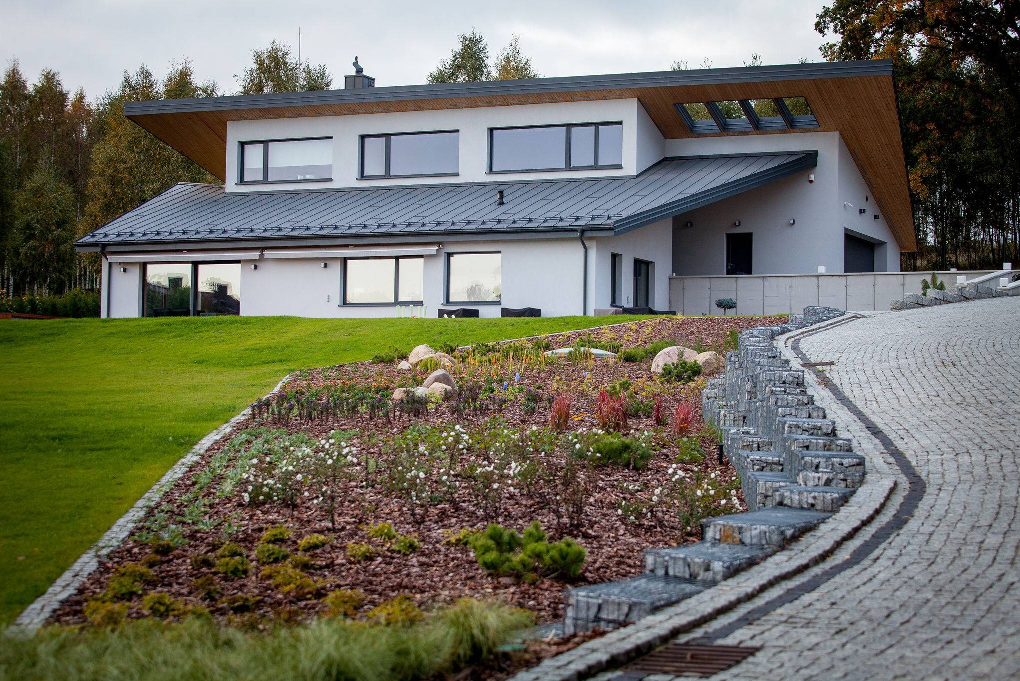Nowoczesny dom jednorodzinny z płaskim dachem, szarą elewacją i dużymi oknami, otoczony zielonym trawnikiem i ozdobnym ogrodem z kamiennymi schodami prowadzącymi do wejścia.