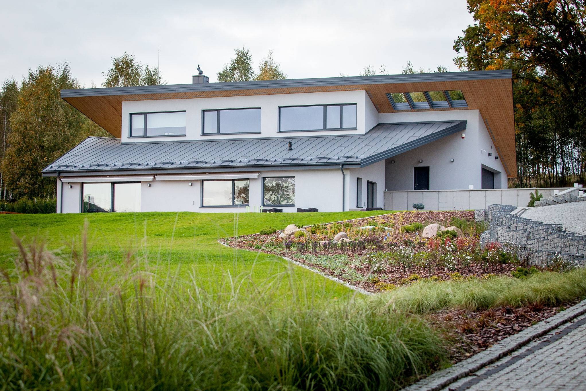 Nowoczesny dom murowany z szarą blachodachówką, drewnianym podbitką i starannie zaprojektowanym ogrodem z kamiennymi elementami.