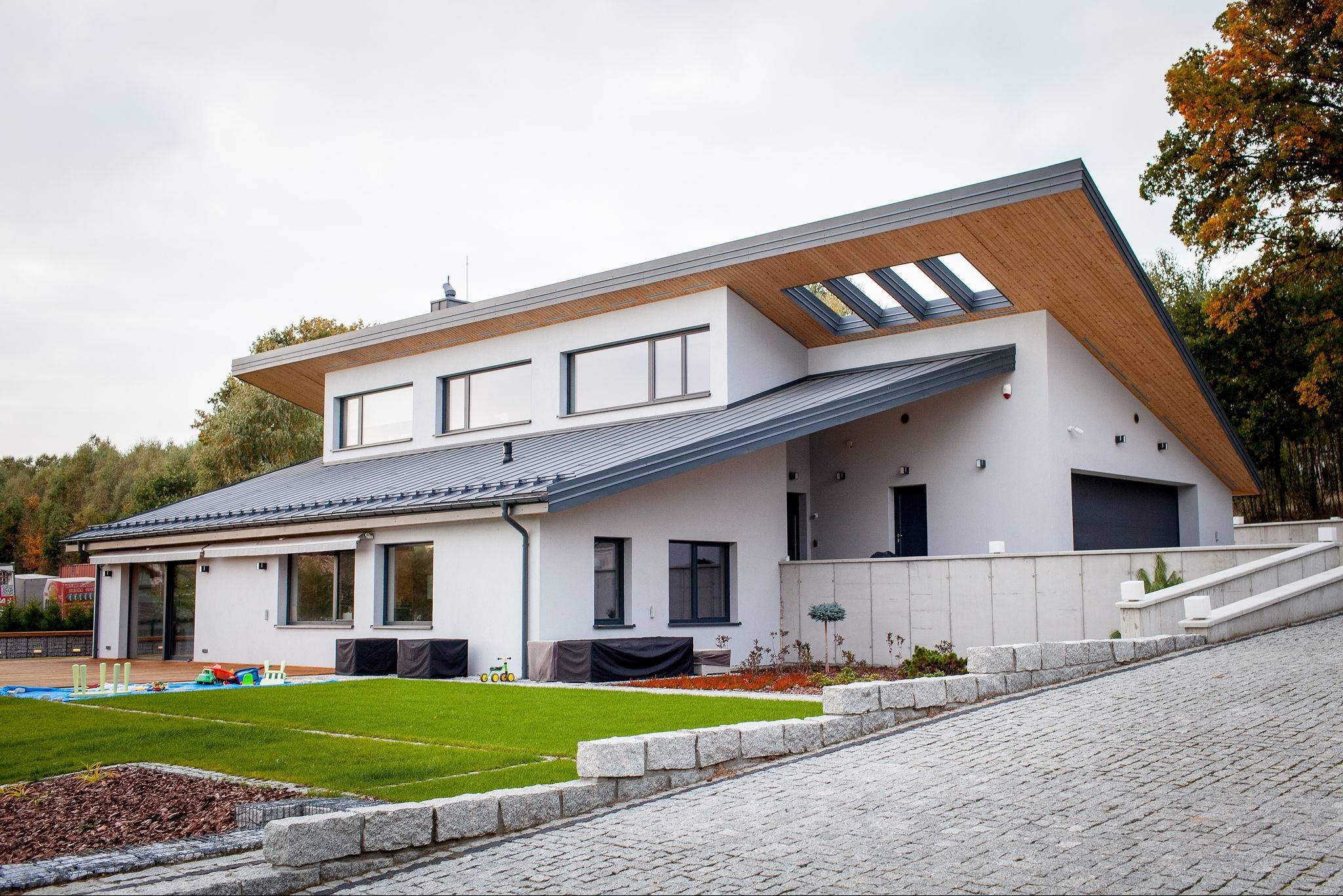 Nowoczesny dom jednorodzinny z płaskim dachem i drewnianym wykończeniem okapu oraz oknami dachowymi, widok z zewnątrz na elewację i zagospodarowany teren z kostką brukową i trawnikiem