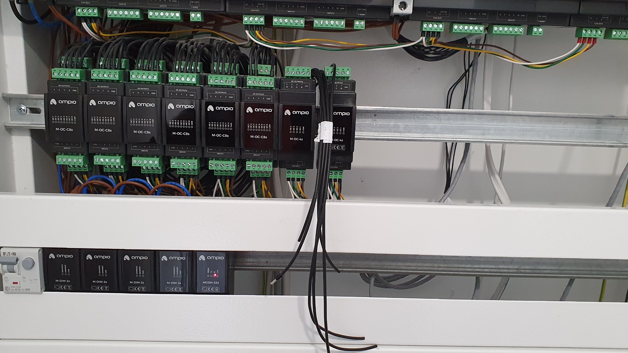 Panel sterowania inteligentnego domu z modułami Ampio M-OC-C8s, M-OC-4s i M-DIM-2s, widoczne liczne kable i złącza, zamontowany na szynie DIN w białej obudowie.