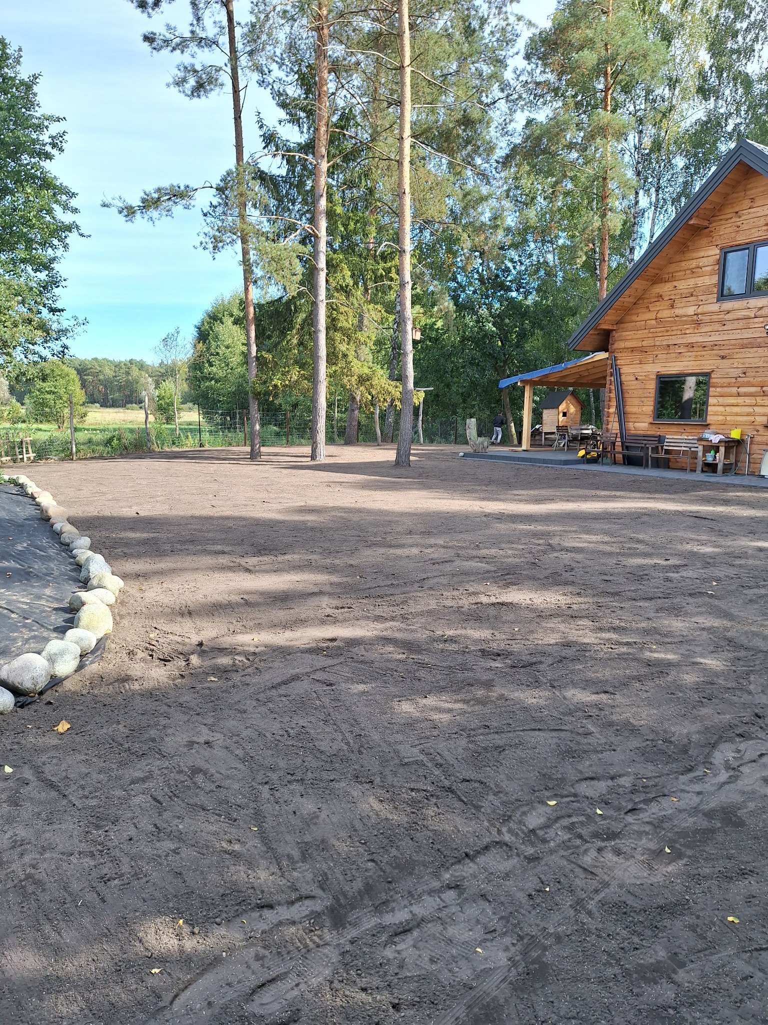 Przygotowany teren pod trawnik z widocznymi śladami po maszynach, obrzeże z kamieni i fragment drewnianego domu w tle.