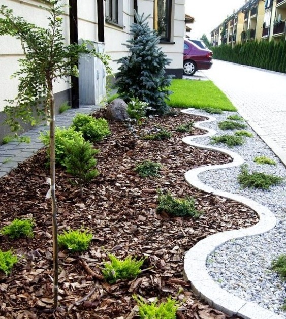 Kaskadowy, wielopoziomowy ogród przydomowy z korą, kamieniami i falującą obwódką, z młodymi drzewkami i krzewami, na tle budynku mieszkalnego i zielonego żywopłotu.