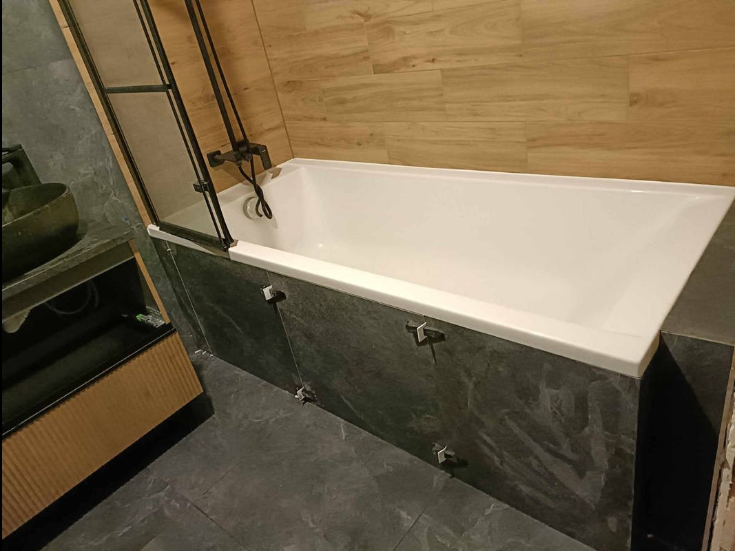 Nowoczesna łazienka z białą wanną obudowaną szarą płytką imitującą kamień, czarną armaturą i drewnopodobnymi płytkami na ścianie w tle.