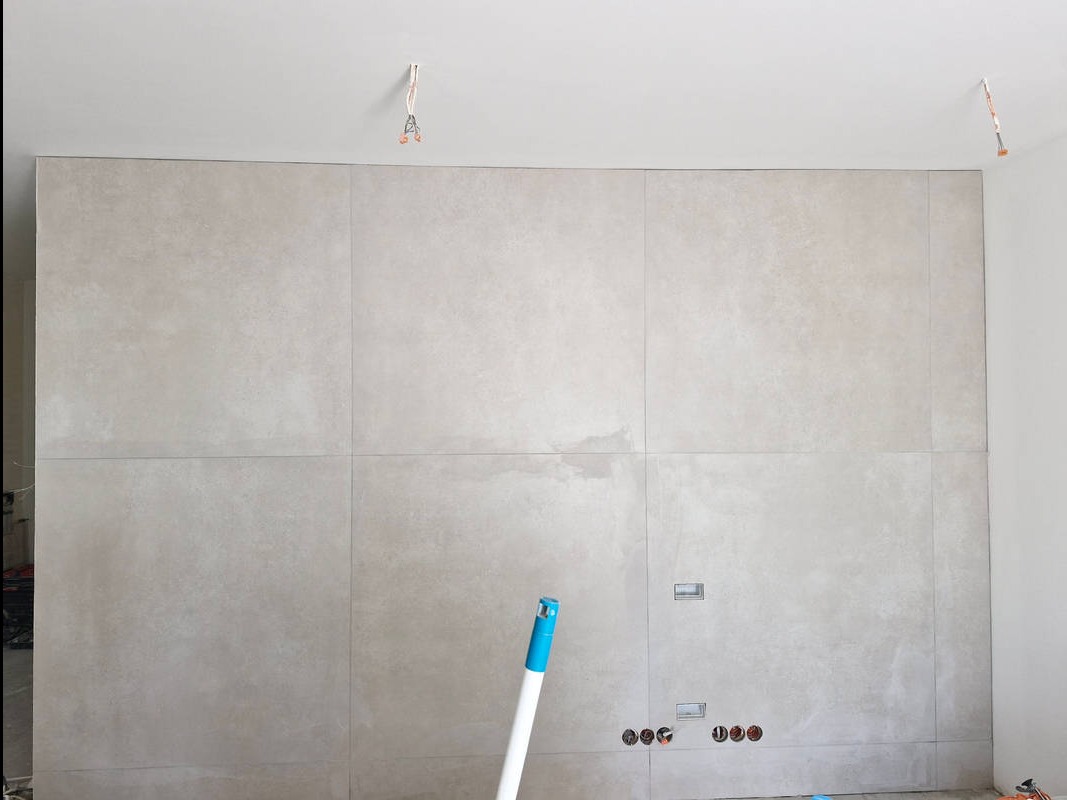 Ściana wyłożona jasnoszarymi, wielkoformatowymi płytkami z widocznymi fugami, niedokończone gniazdka elektryczne, wystające kable z sufitu, biały mop oparty o ścianę.