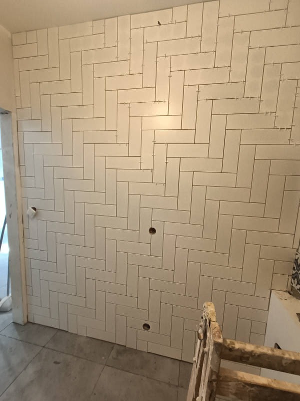 Ściana w trakcie układania białych, prostokątnych płytek w wzór jodełki, widoczne otwory na gniazdka, szpary między płytkami i krzyżyki dystansowe, na pierwszym planie fragment drabiny malarskiej.