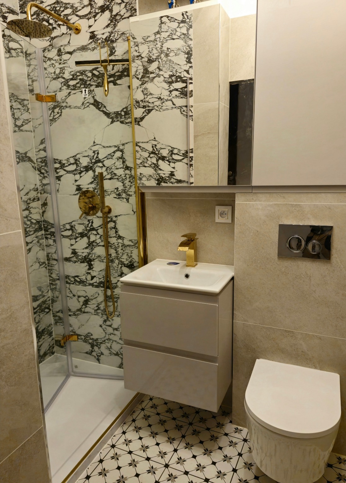 Nowoczesna łazienka z kabiną prysznicową wykończoną marmurem, złotą armaturą i umywalką z szafką w kolorze złamanej bieli, oraz toaletą w tym samym odcieniu, na podłodze wzorzyste płytki.