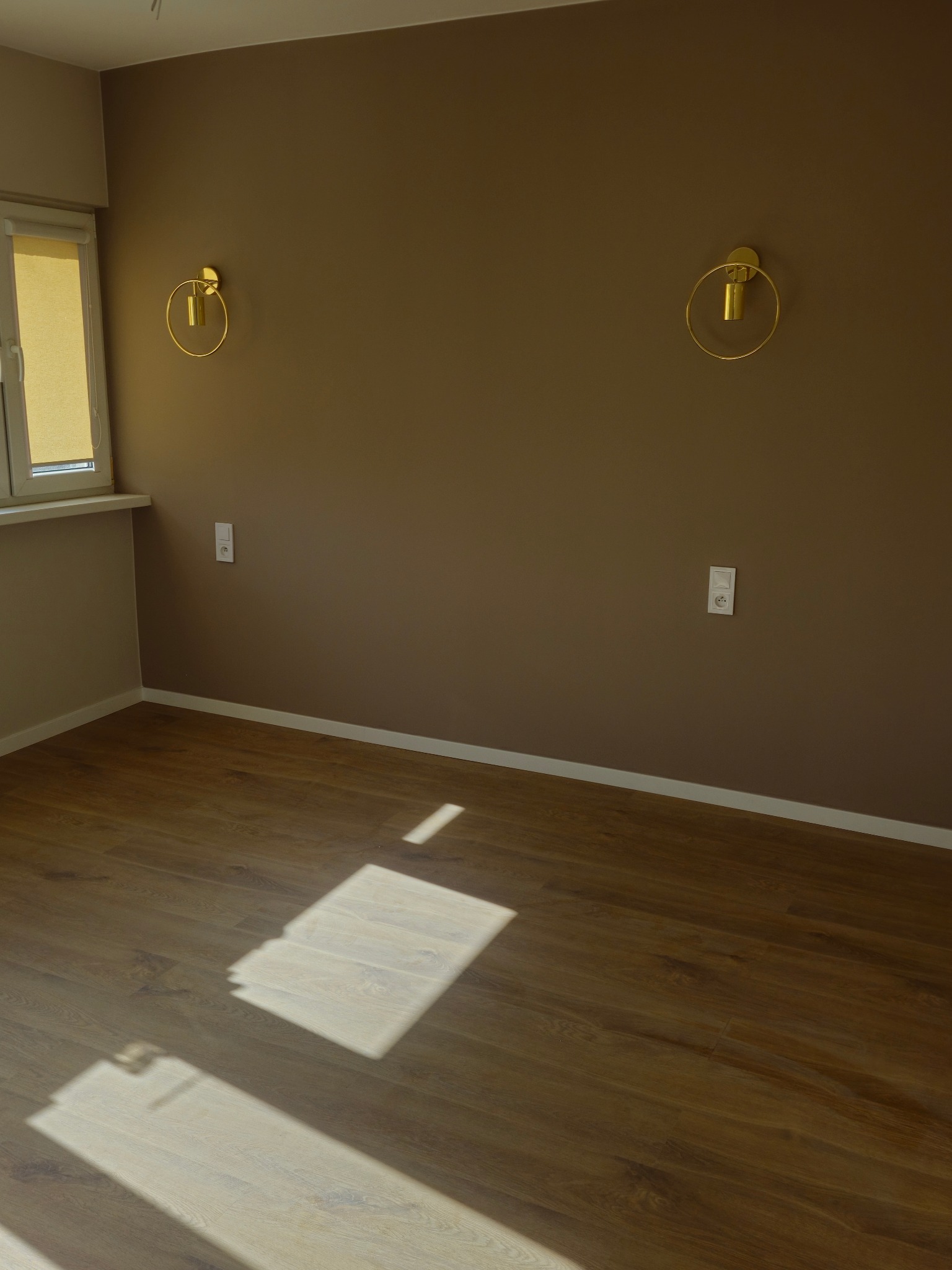 Pokój z brązowymi ścianami, drewnianą podłogą i złotymi lampami ściennymi w kształcie okręgów, oświetlony naturalnym światłem padającym przez okno.