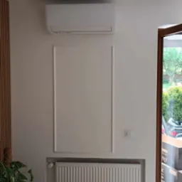 Nowoczesny, biały klimatyzator na ścianie nad grzejnikiem w jasnym pomieszczeniu z drewnianą podłogą i rośliną doniczkową.