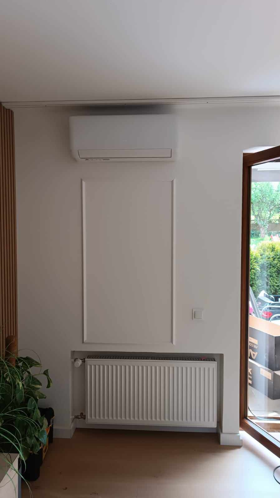 Nowoczesny, biały klimatyzator na ścianie nad grzejnikiem w jasnym pomieszczeniu z drewnianą podłogą i rośliną doniczkową.