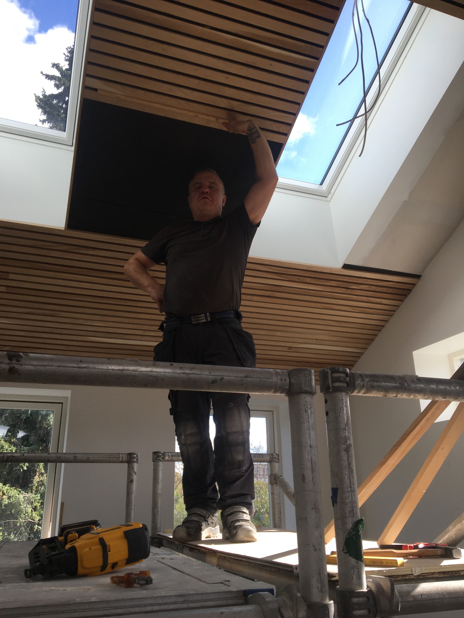 Mężczyzna na rusztowaniu montuje drewniane panele na suficie poddasza, widoczne okno dachowe i narzędzia pracy.