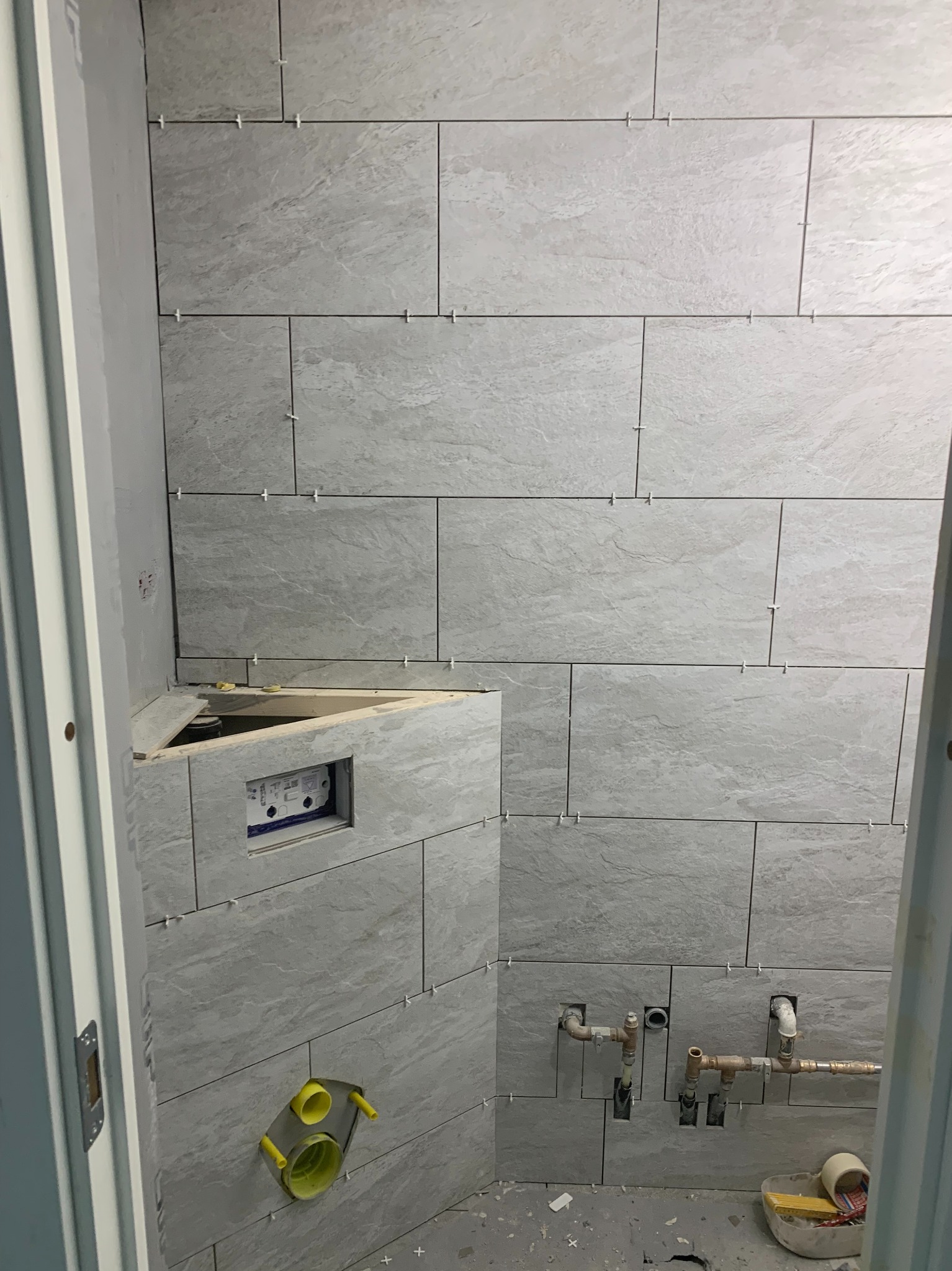 Ściana łazienki w trakcie układania szarych, prostokątnych płytek z użyciem krzyżyków dystansowych, widoczne rury instalacji wodnej i podtynkowy stelaż WC.