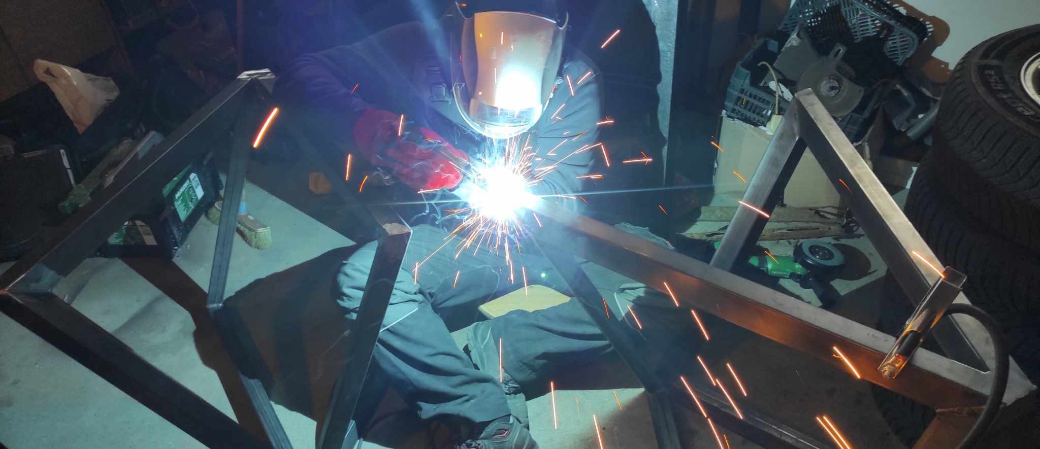 Spawanie metalowej konstrukcji: spawacz w masce i rękawicach łączy elementy, iskry rozświetlają warsztatowe otoczenie z narzędziami i oponą w tle.