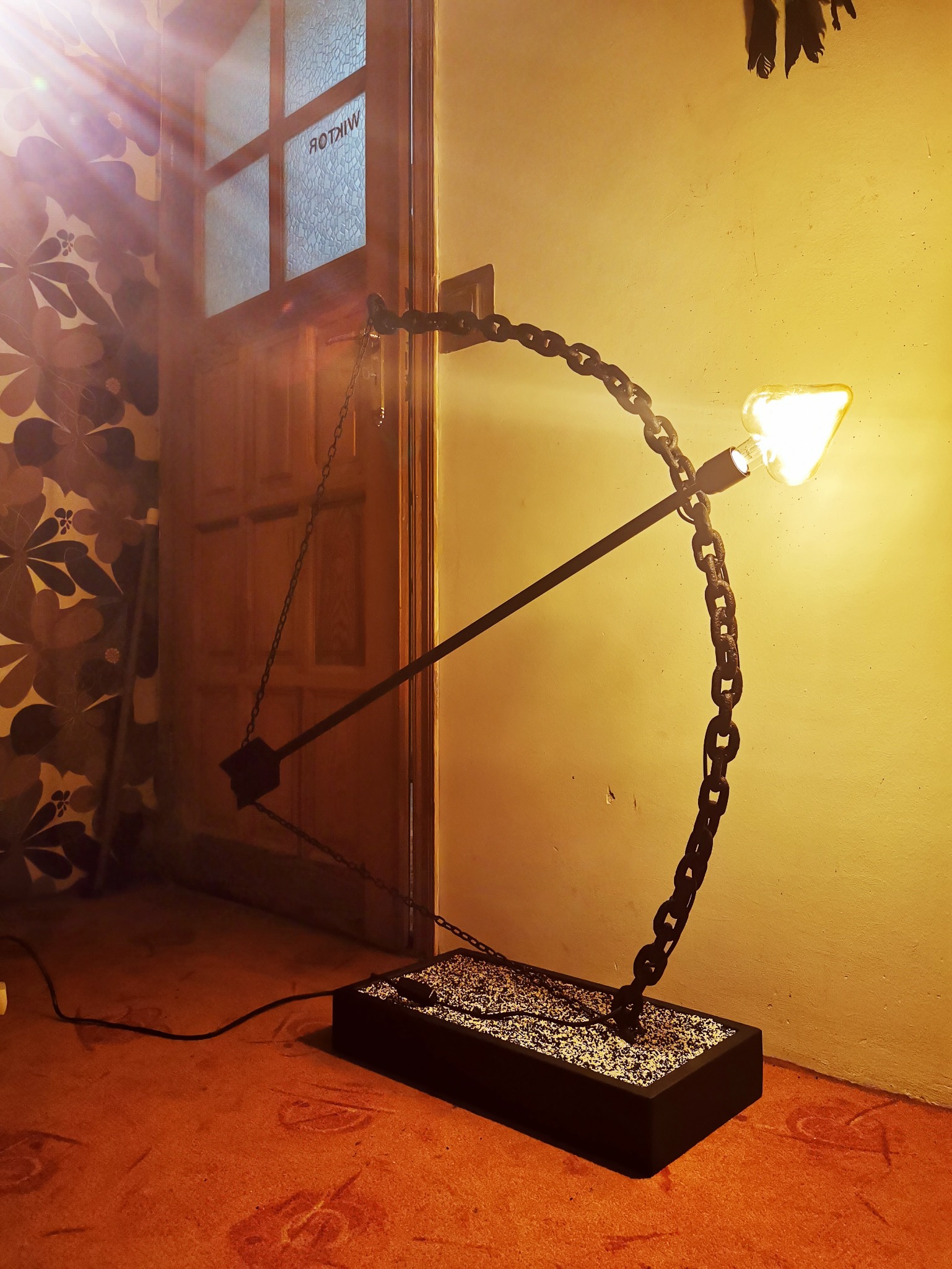 Nietypowa lampa podłogowa w kształcie łuku z łańcucha, z żarówką imitującą strzałę, stojąca na czarnej podstawie wypełnionej białym kruszywem, na tle ściany i drzwi z szybą z napisem 'WIKTOR'.