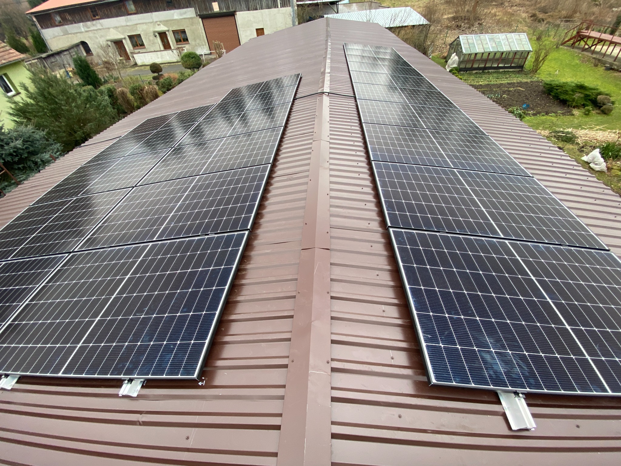 Panele słoneczne zamontowane na brązowym dachu blaszanym, widok z góry, w tle dom i szklarnia.