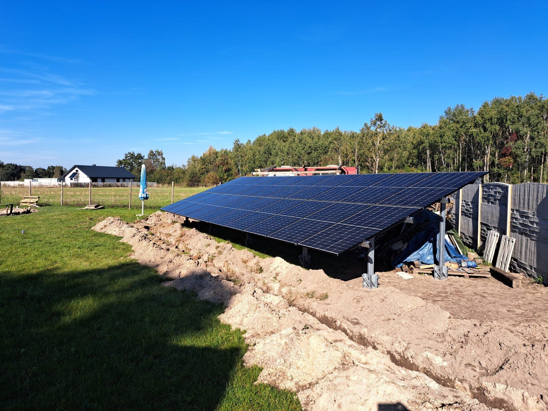 Instalacja paneli słonecznych na metalowej konstrukcji, widoczne wykopane rowy w ziemi, w tle budynek mieszkalny i drzewa pod błękitnym niebem.