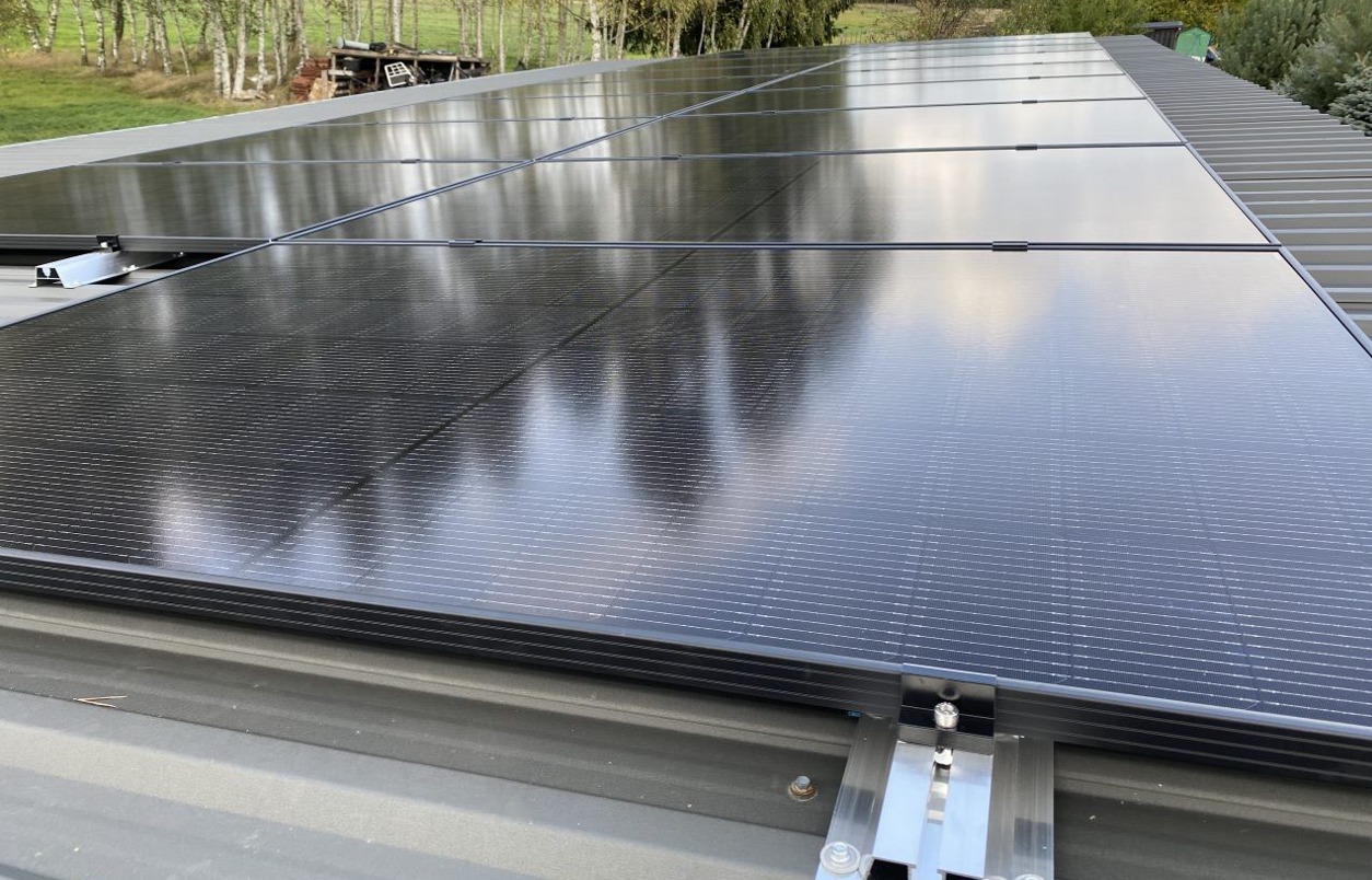Rząd czarnych paneli słonecznych na szarym, metalowym dachu z widocznymi srebrnymi elementami montażowymi, odbijający niebo i drzewa.