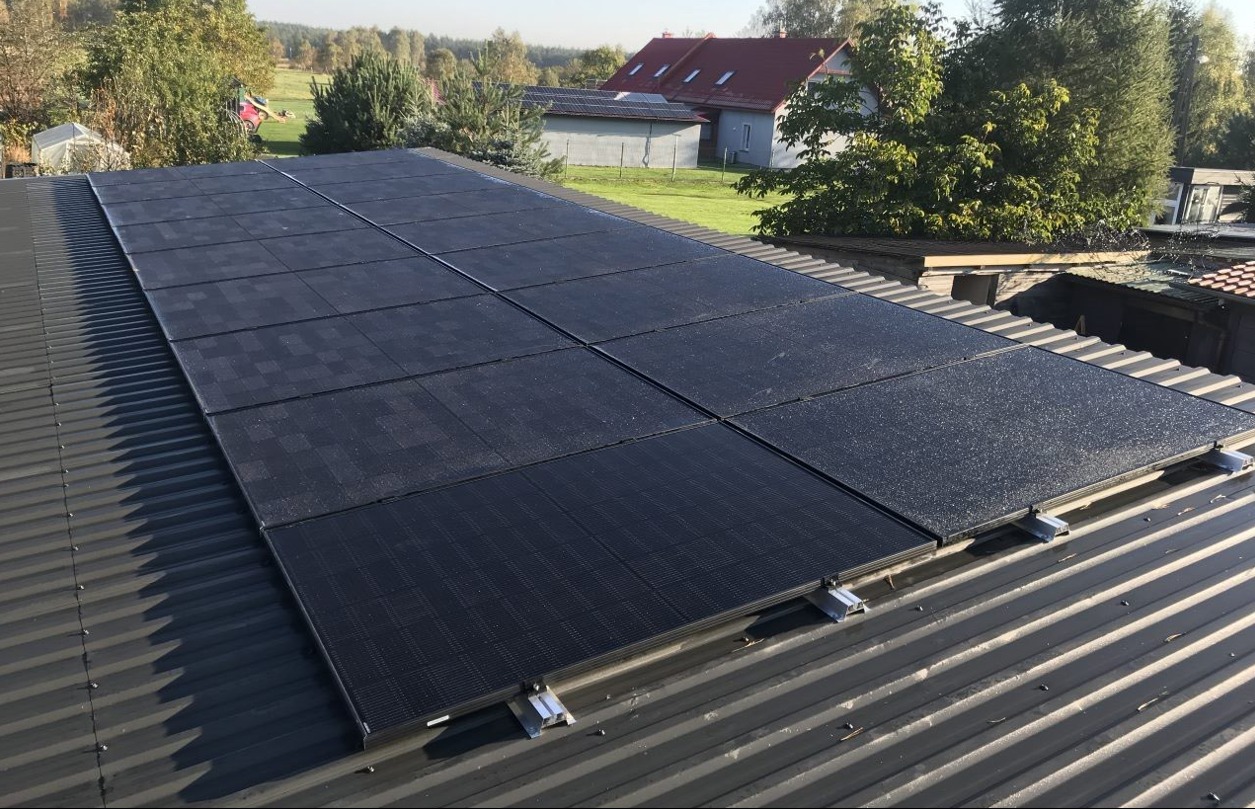 Panel słoneczny na metalowym dachu z widocznymi mocowaniami, w tle dom z czerwoną dachówką i drzewa.