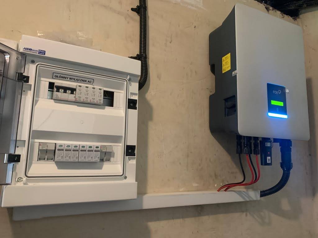 Zamontowany inwerter FoxESS z zieloną diodą LED i otwartą skrzynką z bezpiecznikami AC na ścianie wewnątrz budynku.