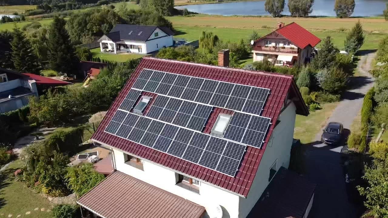 Widok z góry na dom z czerwoną dachówką, na której zamontowane są panele słoneczne, wkomponowane między dwa okna dachowe, w tle jezioro i zabudowa jednorodzinna.