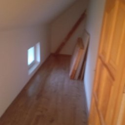 Wykończenia wnętrz - Poddasze z oknem, drewnianą podłogą i drewnianymi drzwiami, w rogu oparte o ścianę płyty.