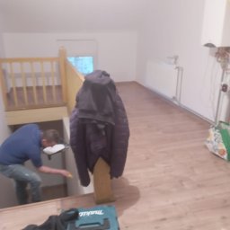 Wykończenia wnętrz - Mężczyzna w niebieskiej koszulce maluje ścianę pod schodami, widoczna drabina, narzędzia i kurtka wisząca na schodach w trakcie prac wykończeniowych.