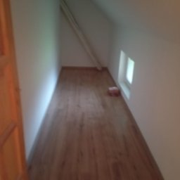 Wykończenia wnętrz - Poddasze z drewnianą podłogą, skośnym sufitem i małym oknem, widoczna rura pod sufitem, niedokończone wnętrze.