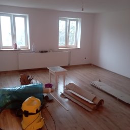 Wykończenia wnętrz - Pokój po malowaniu ścian na biało, z drewnianą podłogą, dwoma oknami, grzejnikiem, małym stolikiem, workiem na śmieci, odkurzaczem i rozłożonymi rolkami papieru zabezpieczającego.