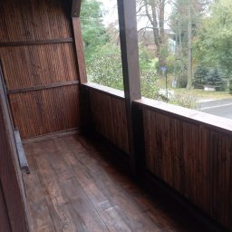 Wykończenia wnętrz - Balkon z drewnianą zabudową, widok na zewnątrz, brązowe deski podłogowe i pionowe panele ścienne, słup podtrzymujący zadaszenie, widoczne drzewa i budynki w tle.