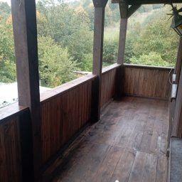 Wykończenia wnętrz - Drewniany balkon z widokiem na zalesione wzgórze, świeżo po impregnacji, z widocznym aplikatorem do impregnatu na podłodze.