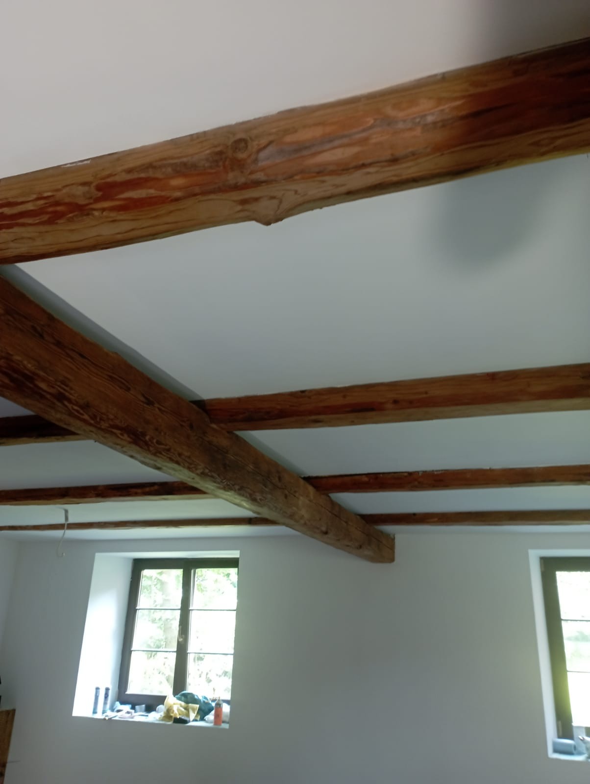 Wnętrze pokoju z pomalowanym na biało sufitem, na którym widoczne są drewniane belki stropowe o wyraźnej strukturze drewna i ciemnobrązowym kolorze. W tle okna z ciemnymi ramami i parapetami,...