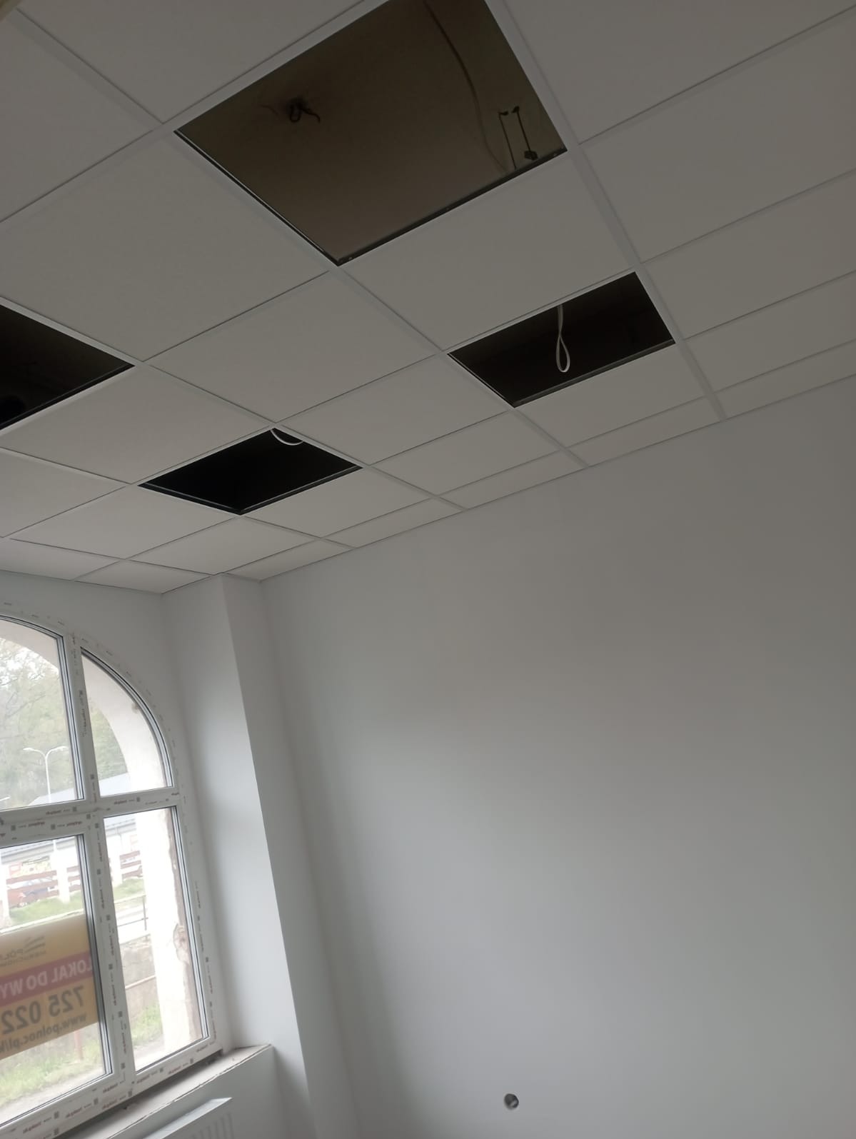 Biały sufit kasetonowy z kilkoma brakującymi panelami, odsłaniającymi instalacje; widok na białą ścianę i okno z łukowym nadprożem w tle.