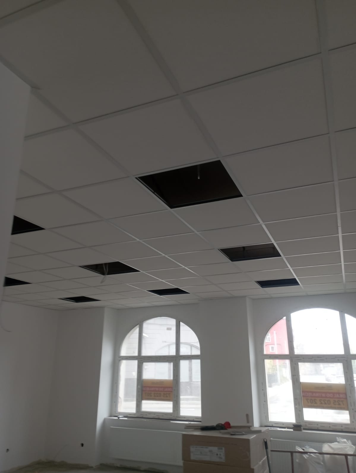 Biały sufit podwieszany z widocznymi otworami na instalacje w jasnym pomieszczeniu z dwoma łukowatymi oknami, widoczne kartony na podłodze.