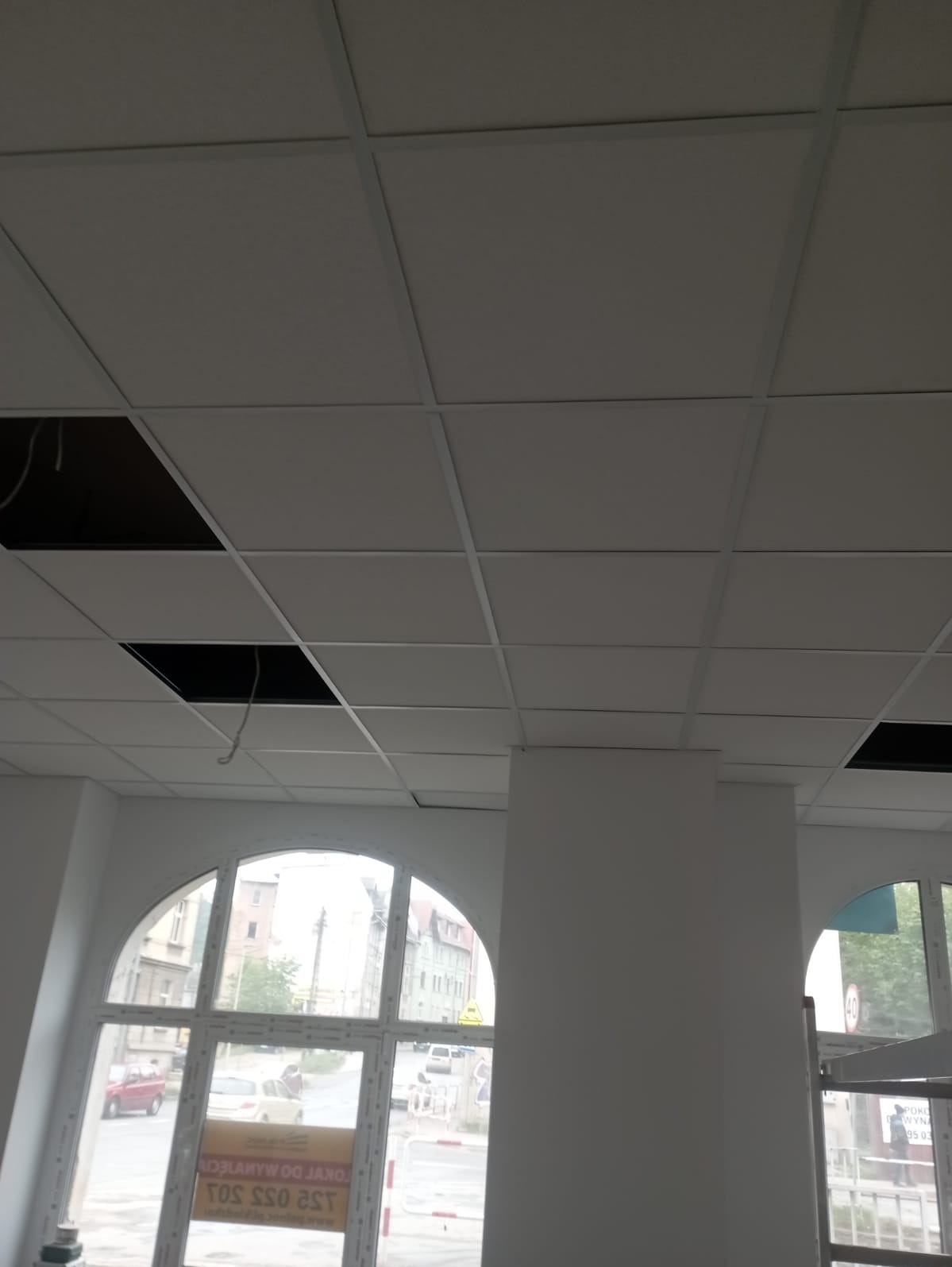 Montaż sufitu podwieszanego kasetonowego w biurze, widoczne otwory na instalacje, białe kasetony i konstrukcja nośna, kolumna konstrukcyjna w tle, okna łukowe z widokiem na ulicę.