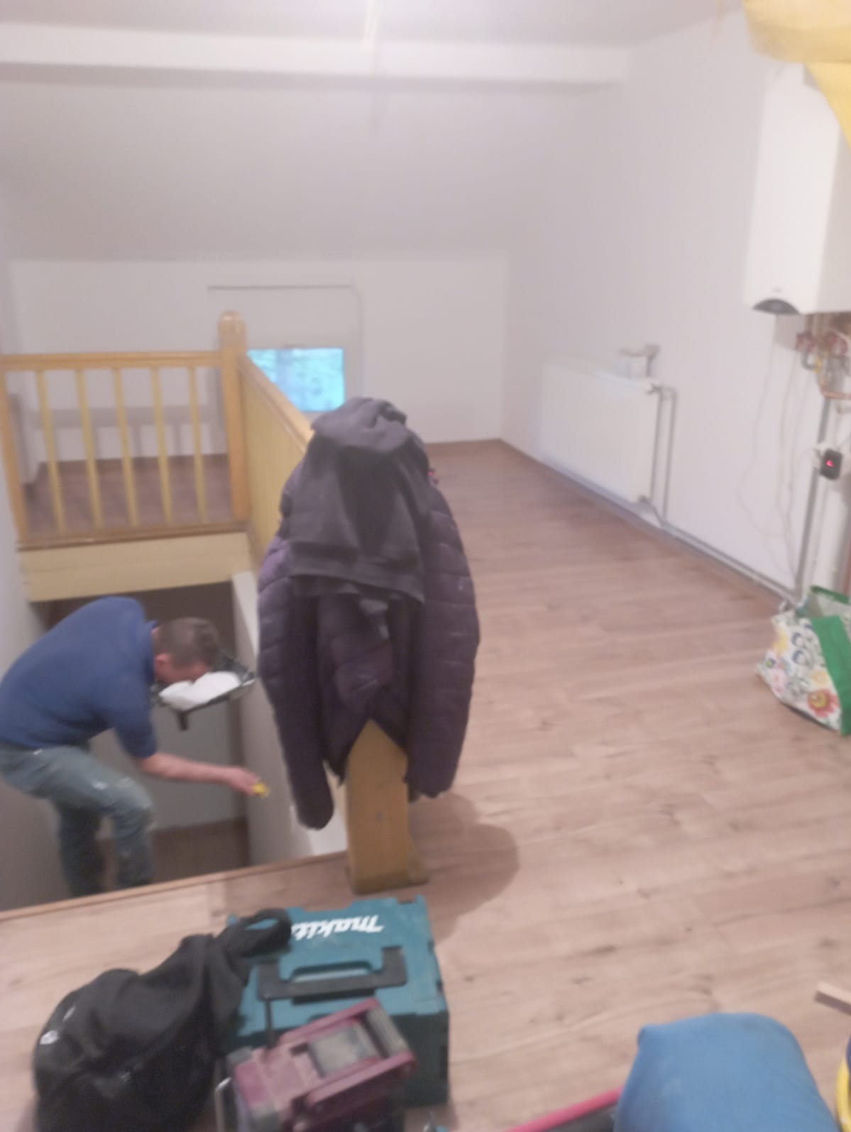 Mężczyzna w niebieskiej koszulce maluje ścianę pod schodami, widoczna drabina, narzędzia i kurtka wisząca na schodach w trakcie prac wykończeniowych.